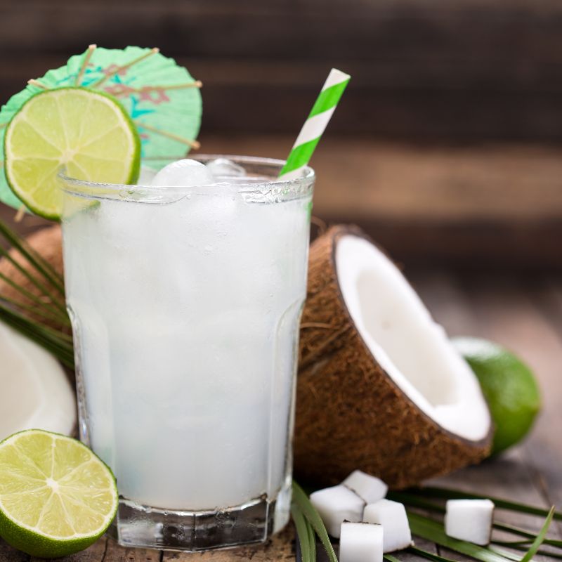 Tropical coconut and lime sparkling drink served over ice in a glass with a green paper straw, surrounded by fresh coconut and lime slices.