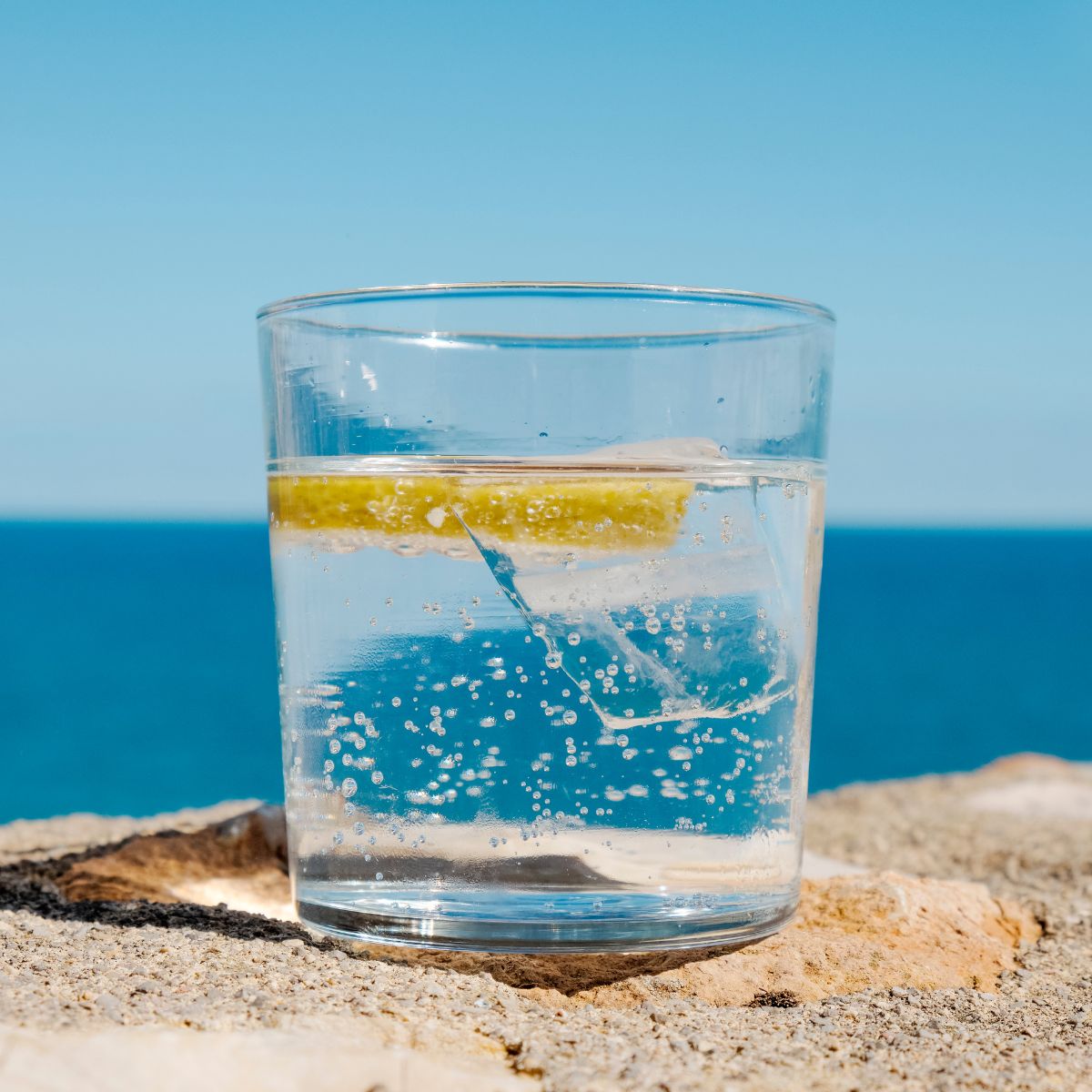 Glass of mountain fall sparkling water with lemon and ice, sitting on a sunlit stone surface by the ocean, under a clear blue sky.